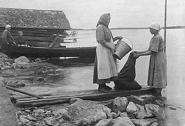Vanhassa kuvassa on kaksi naista rannalla. Toisella on kädessään vesiämpäri, toisella säkki.