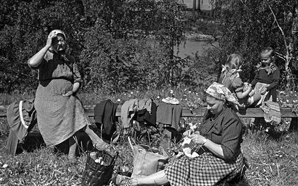 Nainen ja kaksi naista istuskelevat puisella kaiteella, yksi nainen istuu nurmikolla kutomassa. Kaiteella on henkilöiden takkeja ja maassa eväskassi.